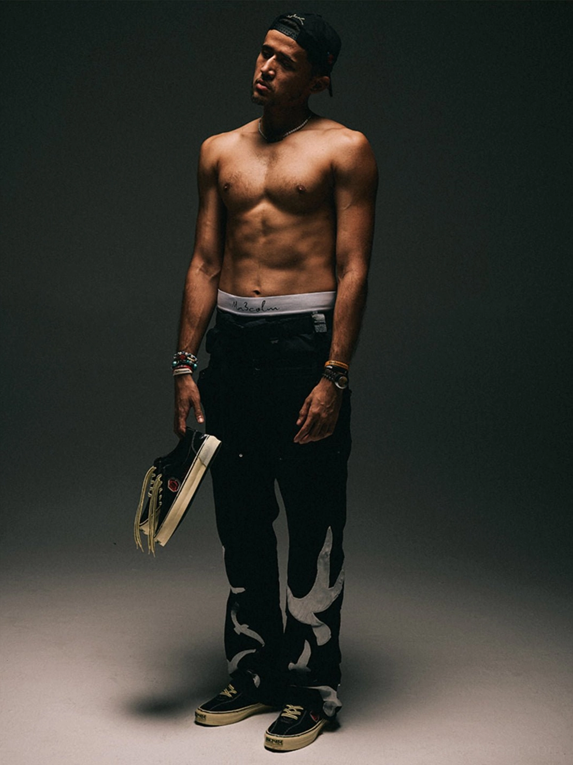 Full-body male streetwear model holding and wearing black embroidered low-top skate shoes with graphic black trousers