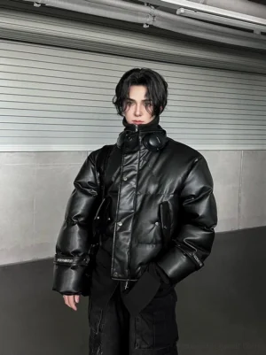 Front half-body view of a black faux leather puffer jacket with matte shell, high stand collar, snap fasteners, padded construction, and adjustable cuff straps in an industrial concrete hallway