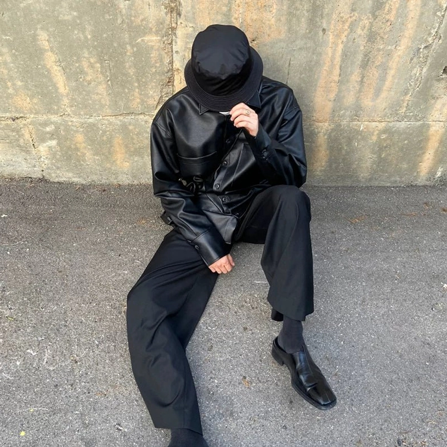 Male model wearing a black faux leather shirt jacket in a relaxed oversized Seoul streetwear outfit with bucket hat, wide leg trousers and leather loafers