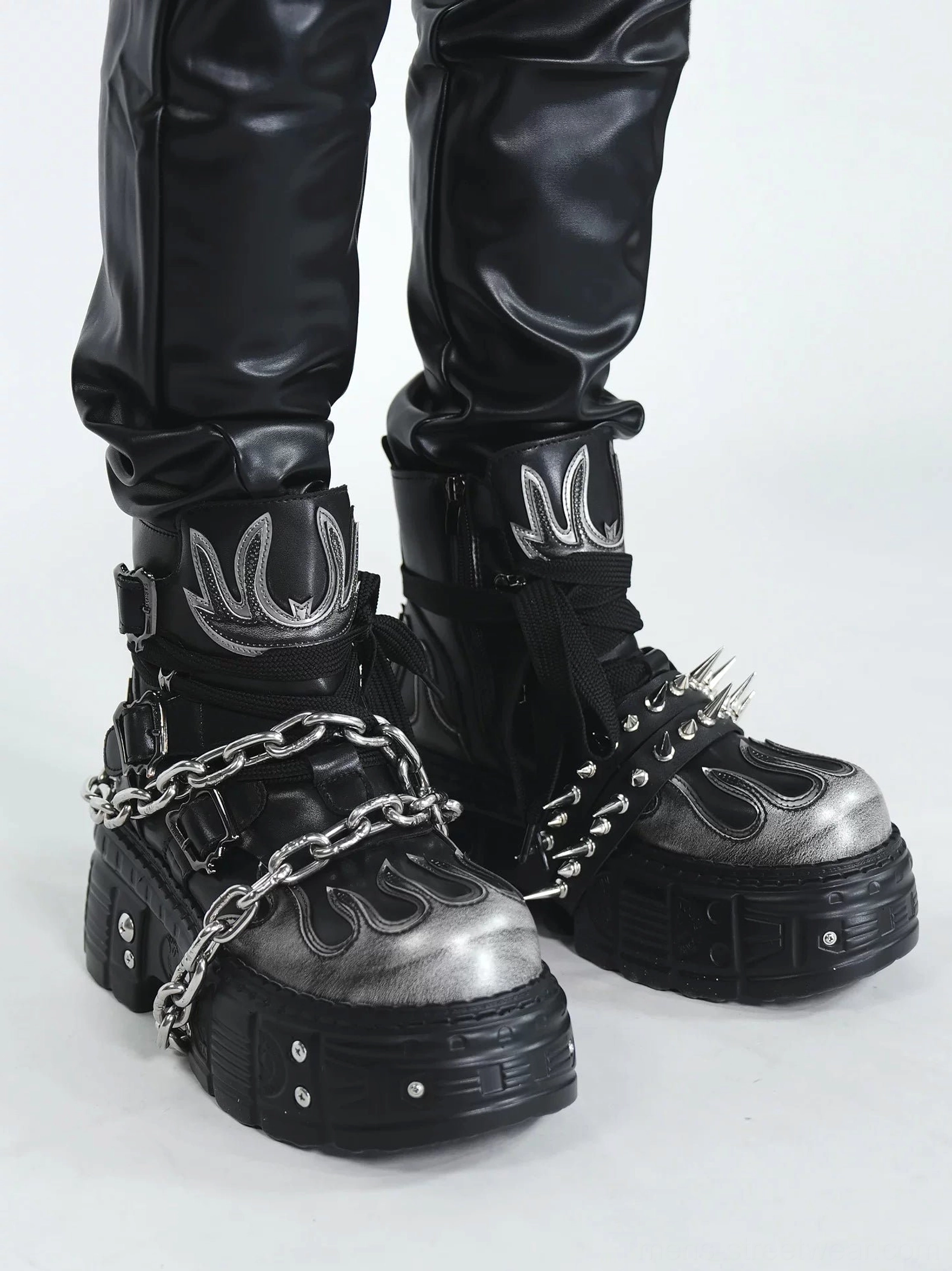 Low angle view of black and silver flame chain spike platform ankle boots styled with black faux leather trousers and chunky lug soles