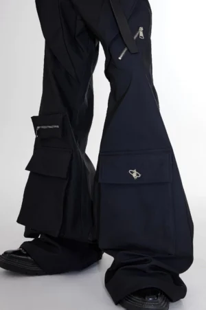 Front close-up of black flared cargo trousers showing oversized flap cargo pockets, side zipper pockets, metal hardware, and matte black fabric