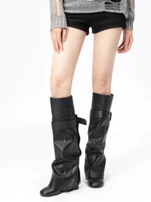 Model wearing black foldover knee-high streetwear boots with slouchy shafts and buckle straps styled with black shorts and a gray knit top