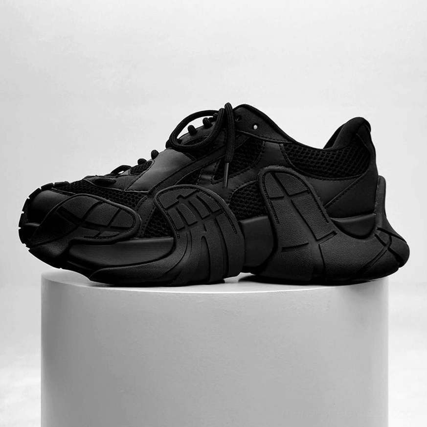 Black futuristic chunky platform sneakers displayed on a studio pedestal, showing lace-up construction and distinctive segmented techwear outsole