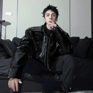 Male model seated in black croc embossed faux leather moto jacket showing zipper front and oversized structured shoulders