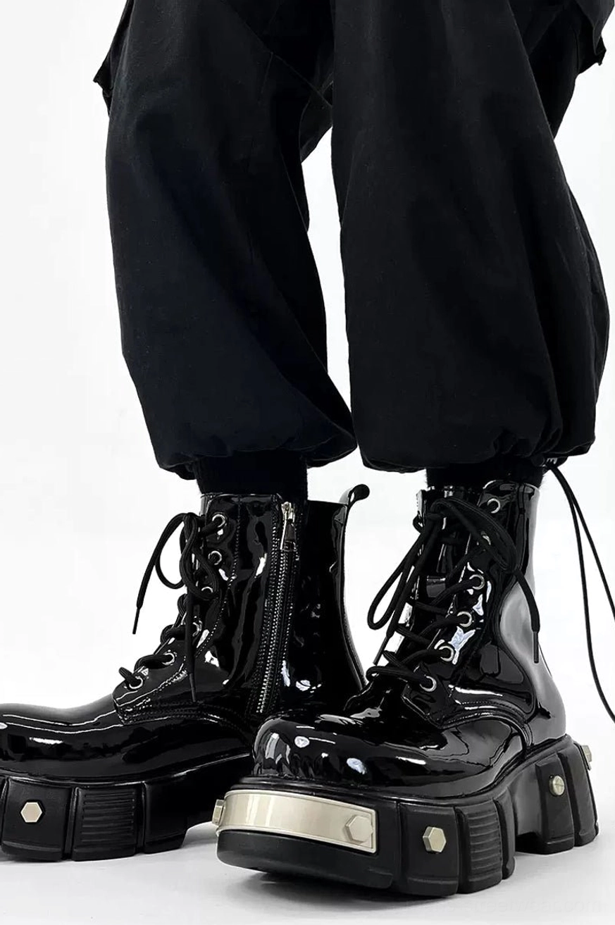 Front view of black glossy platform combat boots worn with black cargo pants, featuring lace-up uppers, silver toe plate, and hexagonal rivets