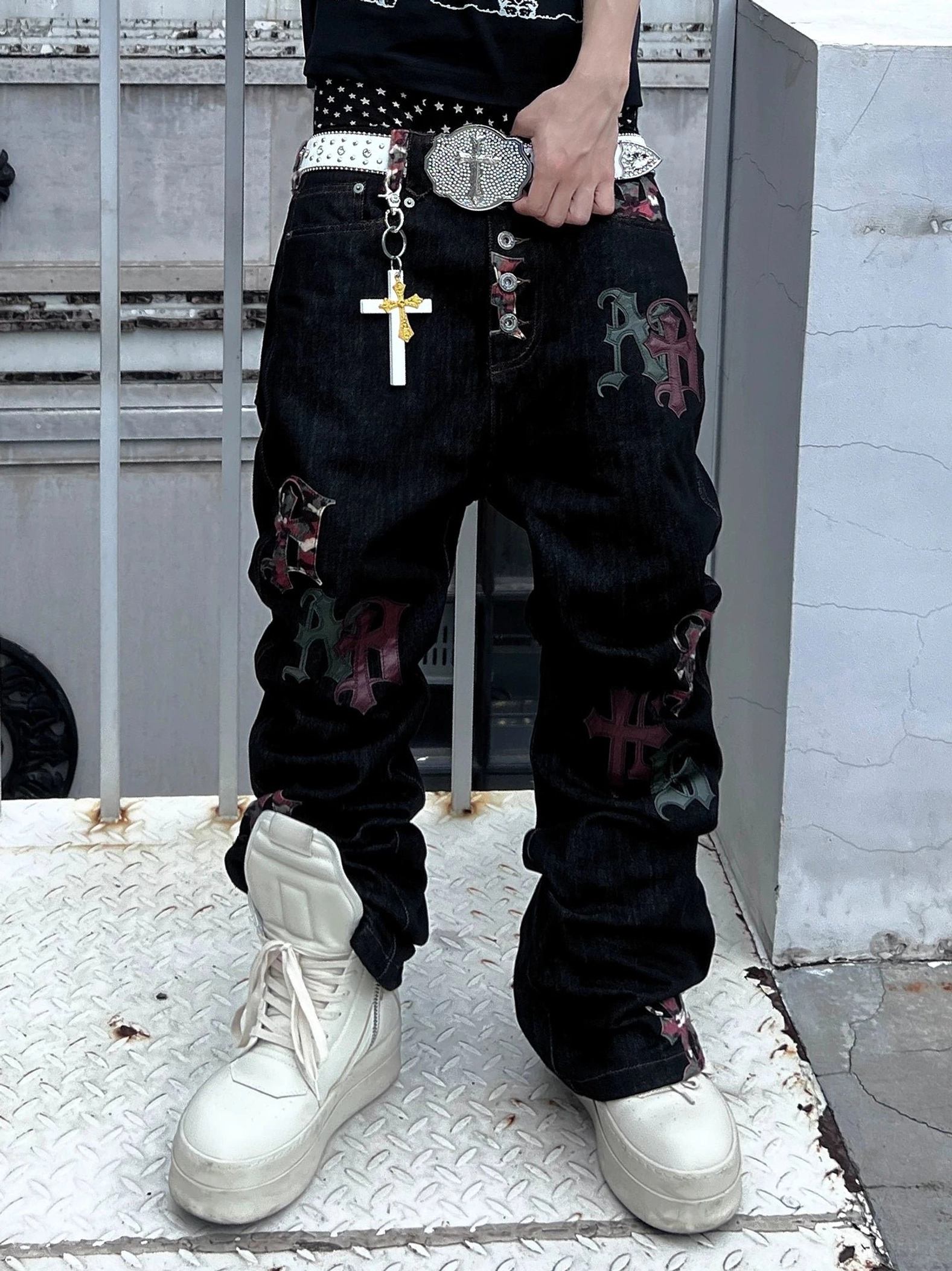 Close-up of black Gothic appliqué denim jeans showing cross patches multi-button fly belt accessory and white platform sneakers