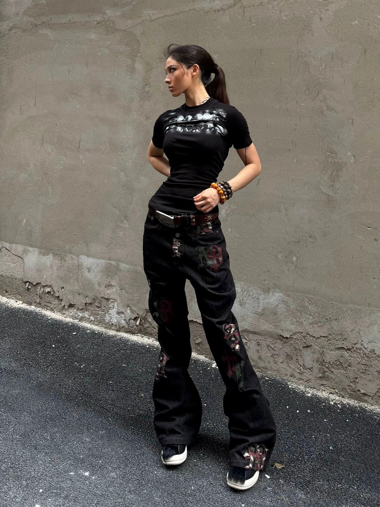 Female model styling black Gothic letter appliqué relaxed straight-leg denim jeans against an urban concrete wall