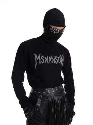 Three quarter view of a black gothic logo mock neck long sleeve streetwear top styled with futuristic sunglasses leather gloves studded belt and dark avant garde trousers