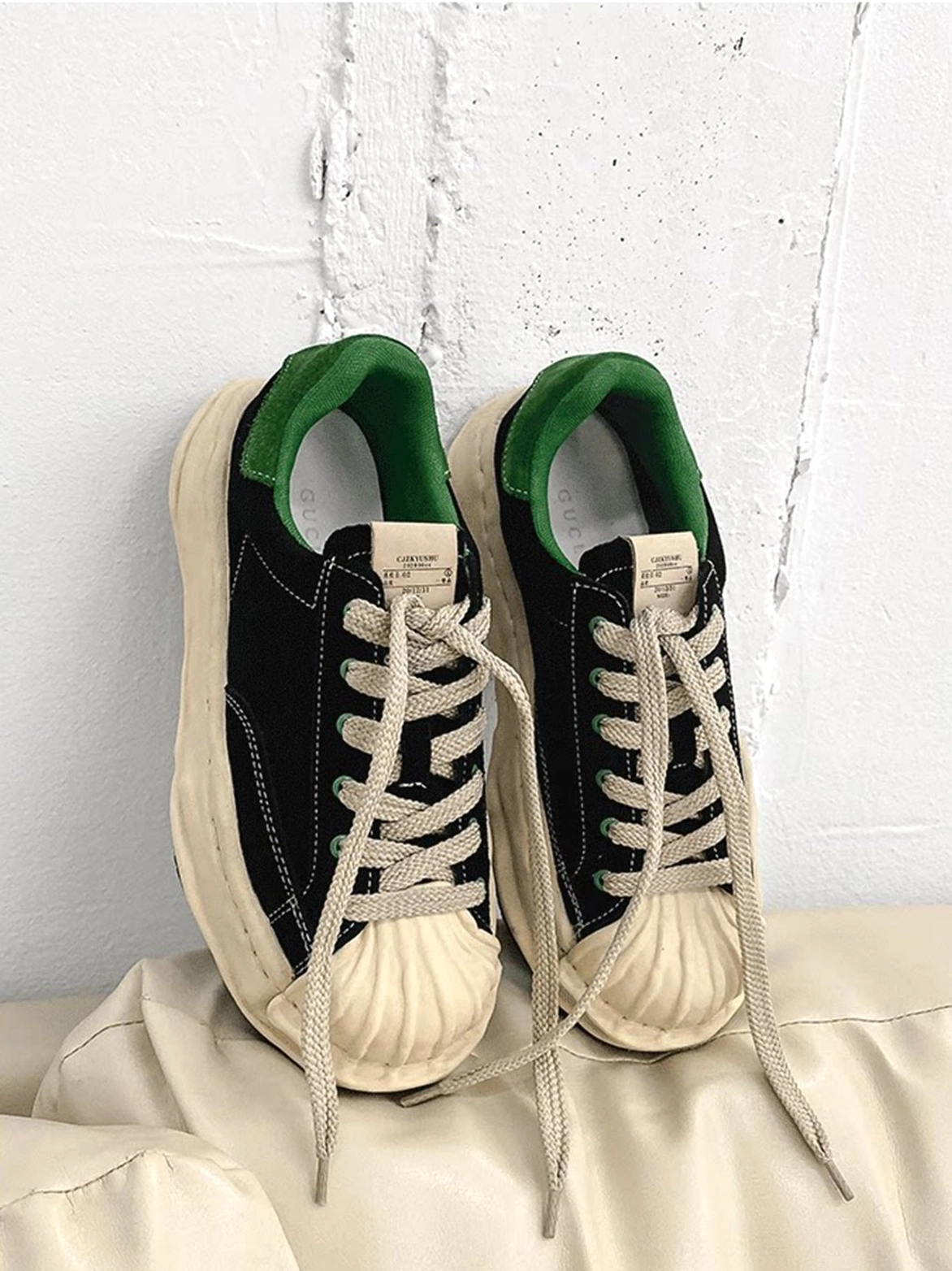 Black and green shell-toe platform sneakers displayed on a leather sofa with visible green lining, cotton-style laces, and thick cream midsole
