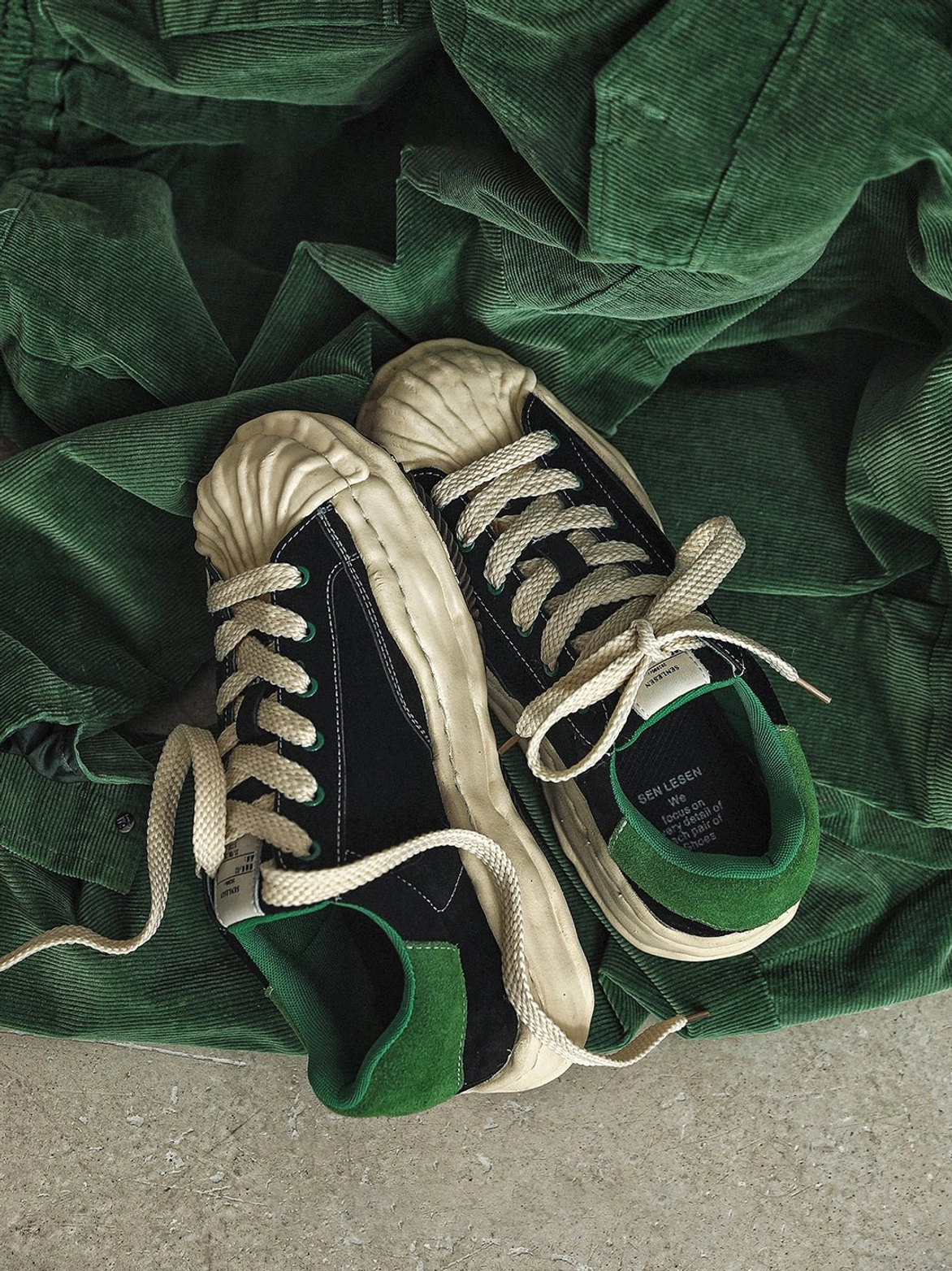 Black shell-toe platform sneakers with green accents shown on dark green corduroy fabric, highlighting suede texture, laces, and heel tab