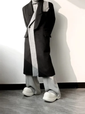 Low-angle detail of the long black wool blend overcoat showing the draped grey front panel, notch lapel, layered collar, chest welt pocket, and long hem