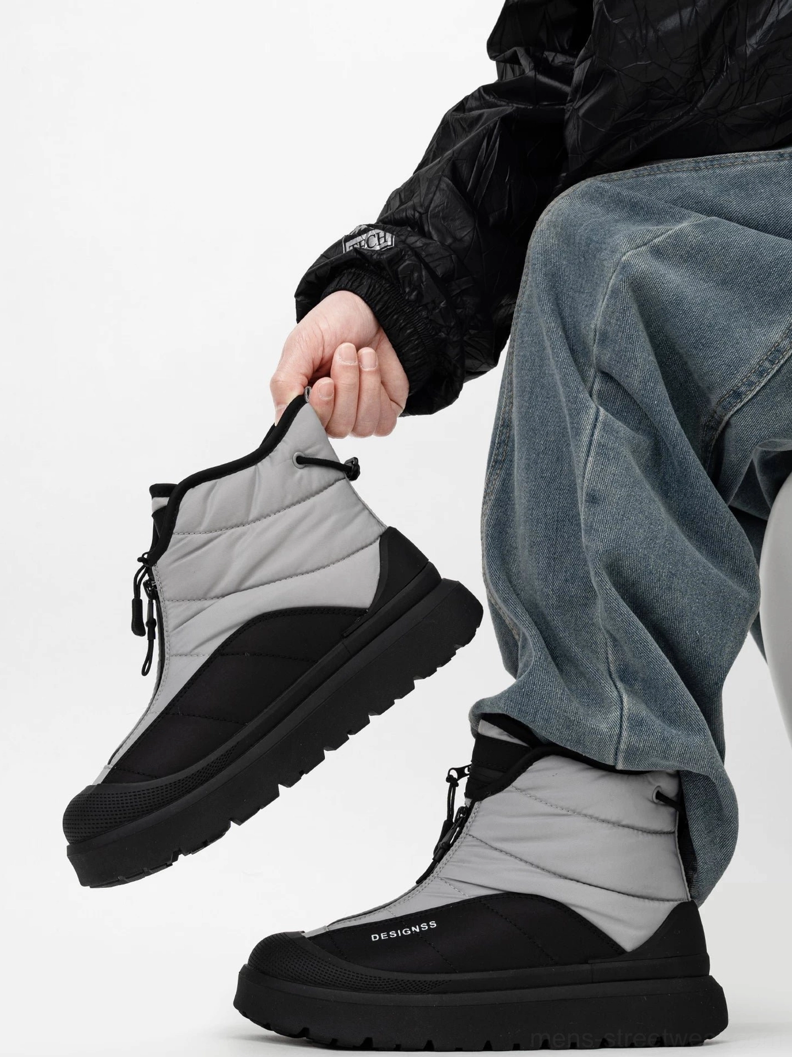 Side profile of a black and grey mid-calf winter snow boot worn with blue jeans, featuring front zipper and rear pull tab