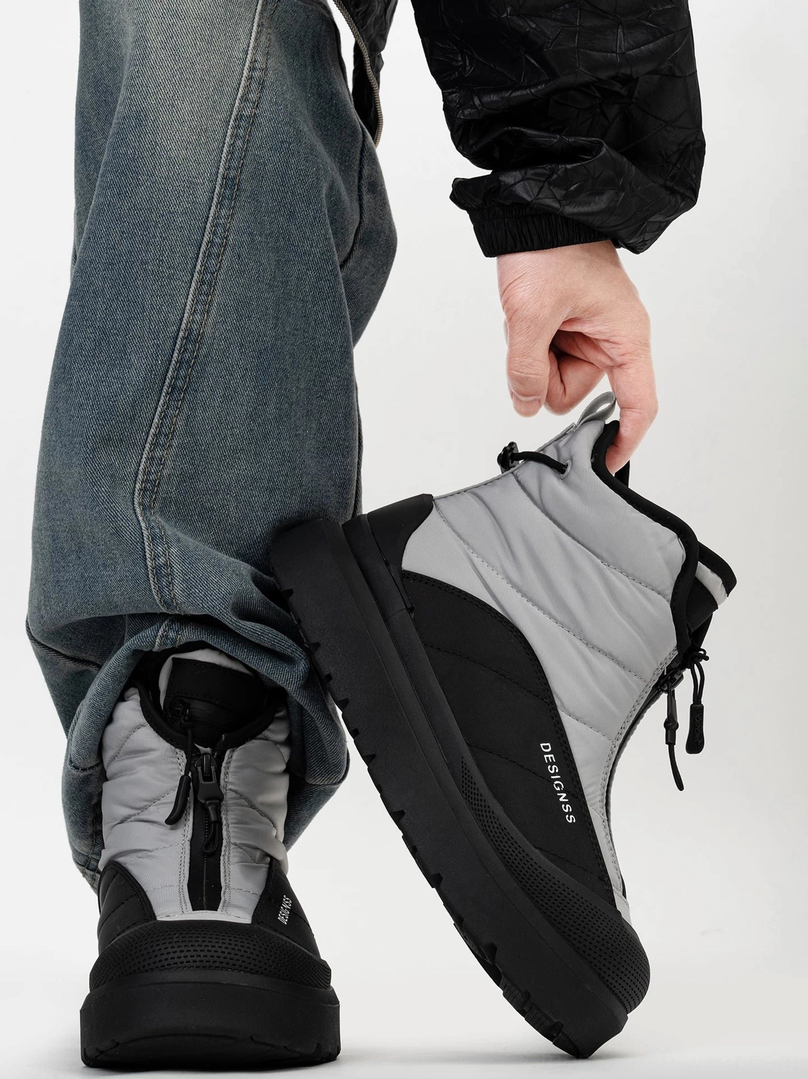 Black and grey quilted zip-up snow boot being fitted onto a foot, showing adjustable drawcord collar and padded mid-calf construction