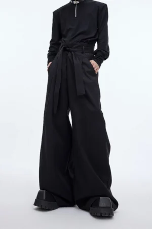 Black high-waist draped wide-leg streetwear trousers with attached waist sash styled with black half-zip top and platform boots