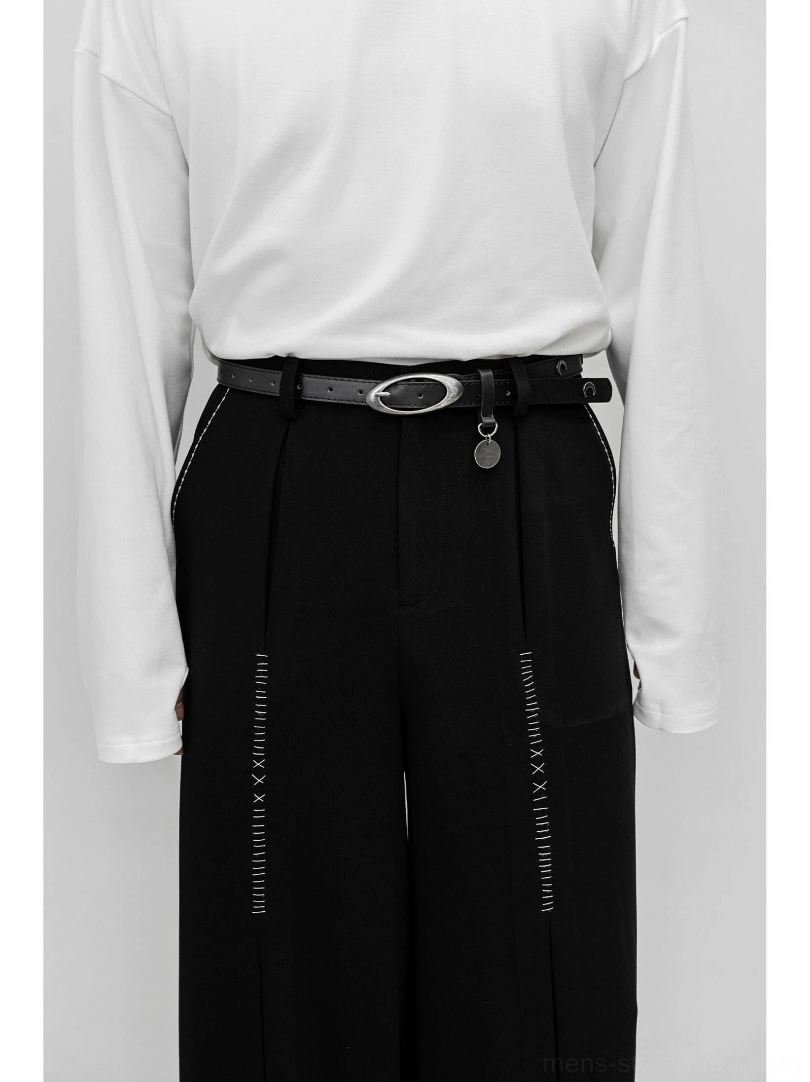 Close-up of black high-waisted wide-leg trousers showing front pleats, waistband construction, decorative leg stitching, and oval metal belt buckle