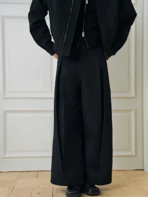 Front low-angle view of black high-waisted wide-leg pleated trousers styled with a cropped black zip jacket in a minimalist indoor studio