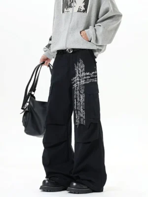 Black ink script cargo pants shown from the front with a relaxed wide-leg fit, large utility pockets, white cross-script graphic, gray zip-up hoodie, tote bag, and leather shoes