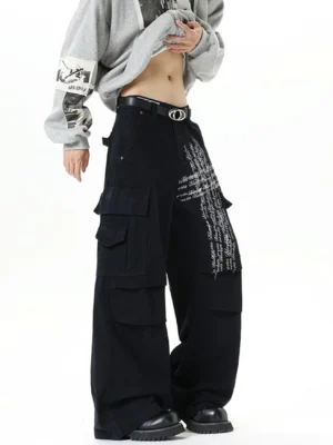 Black ink script wide-leg cargo pants shown from a low front three-quarter view with oversized flap pockets, white graphic lettering, gray hoodie styling, double-ring belt, and black leather shoes