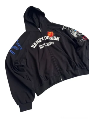 Oblique front view of black Korean streetwear zip hoodie showing oversized cropped fit, metal zipper, varsity chest graphic, and detailed sleeve embroidery