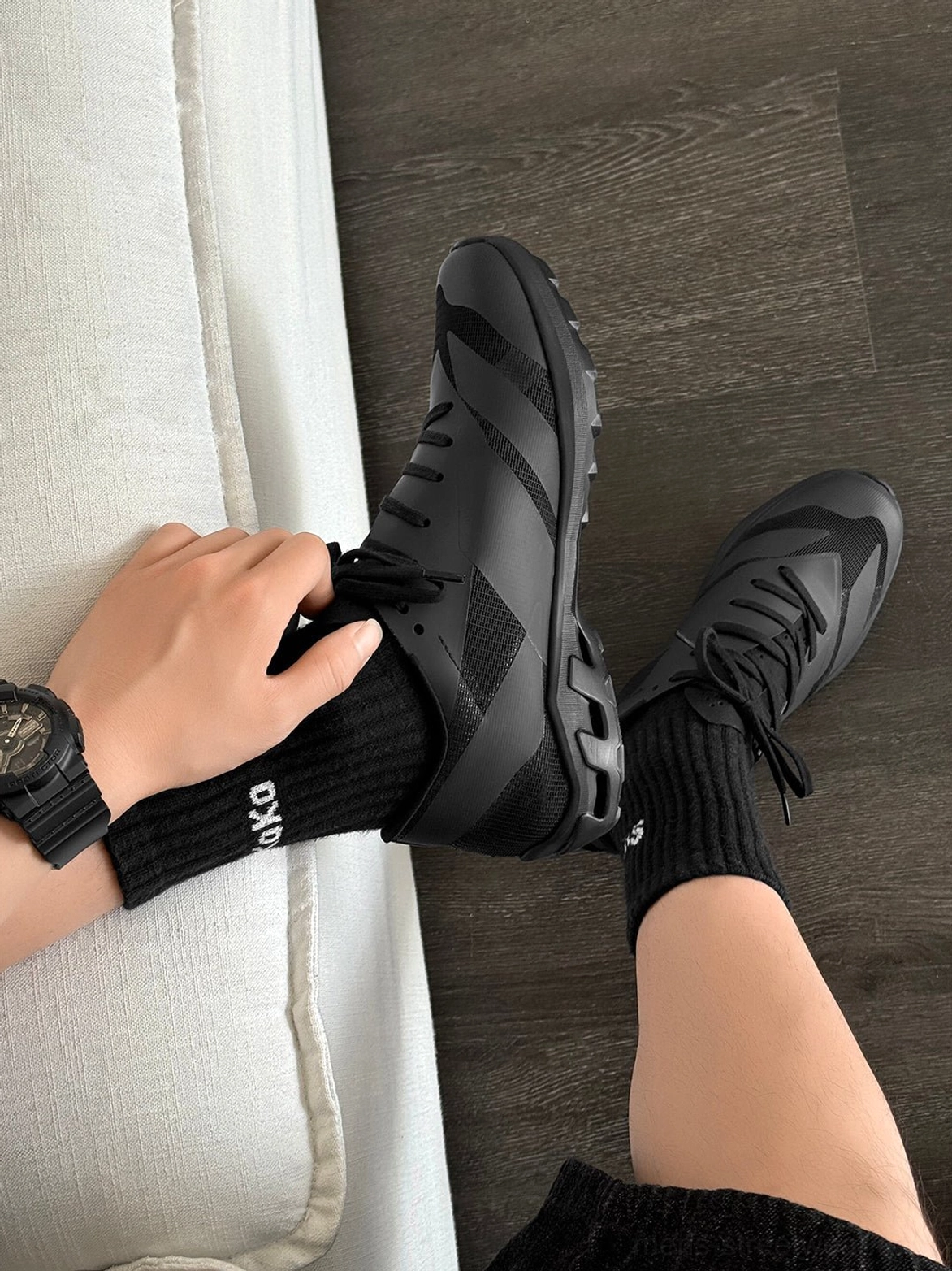 Person putting on a black Korean streetwear sneaker beside a sofa, highlighting the low-cut silhouette, tonal laces, and everyday casual styling