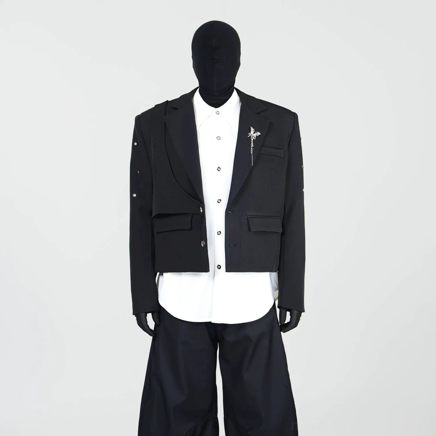 Front studio display of a black layered cropped blazer with double lapels, metallic brooch pin, sleeve studs, and contemporary streetwear tailoring