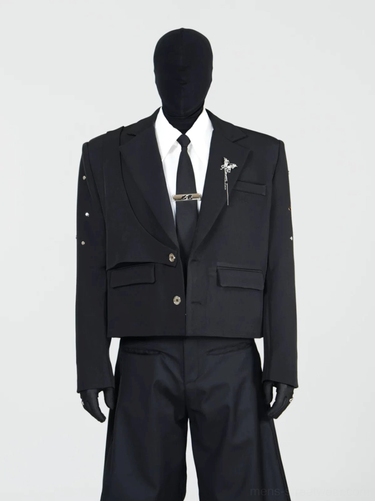Front view of a black cropped layered blazer with double flap pockets, metal brooch, sleeve stud details, and tailored streetwear silhouette