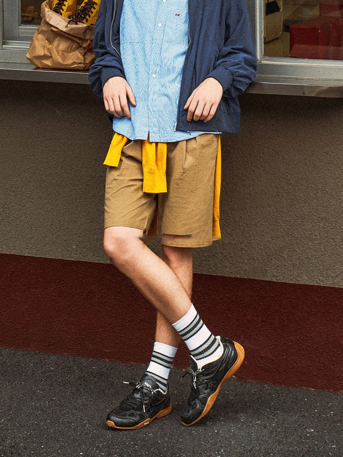 Male model styling black layered streetwear runner sneakers with shorts and a yellow fabric accent in an outdoor storefront setting