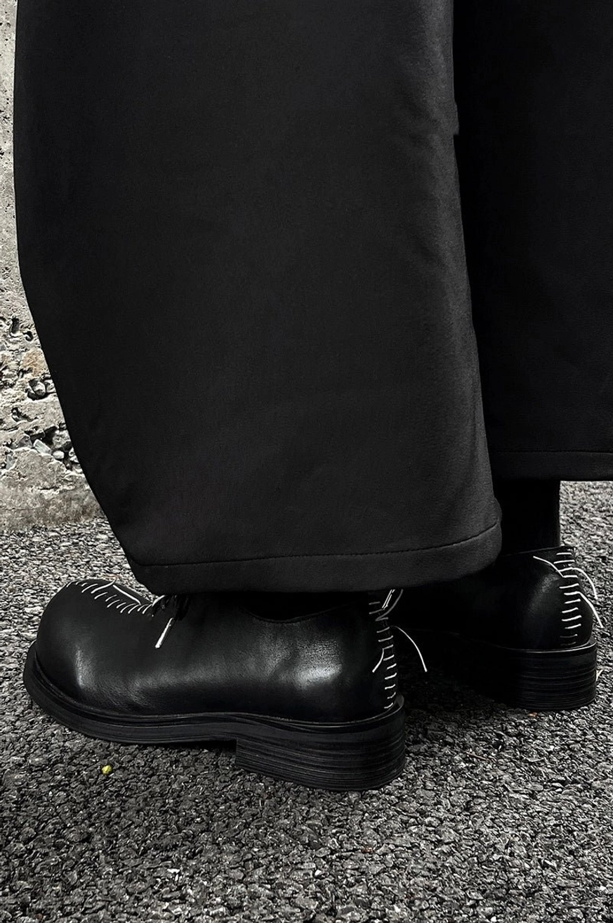 Black leather contrast stitch Oxford shoes worn with wide-leg black trousers in a Korean streetwear outfit on urban pavement