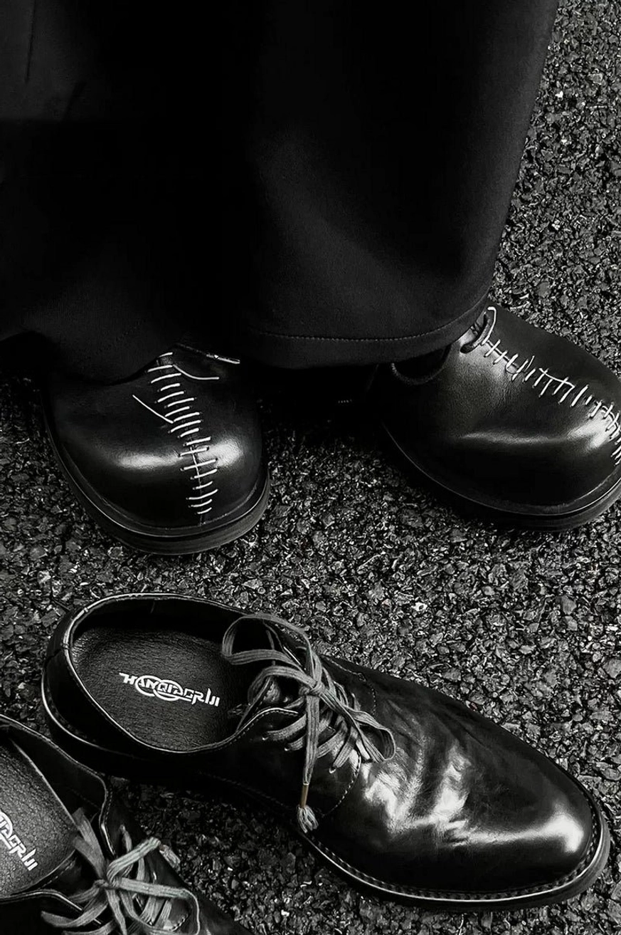 Black polished leather Oxford shoes with white decorative toe stitching displayed as an overhead flat lay on asphalt pavement