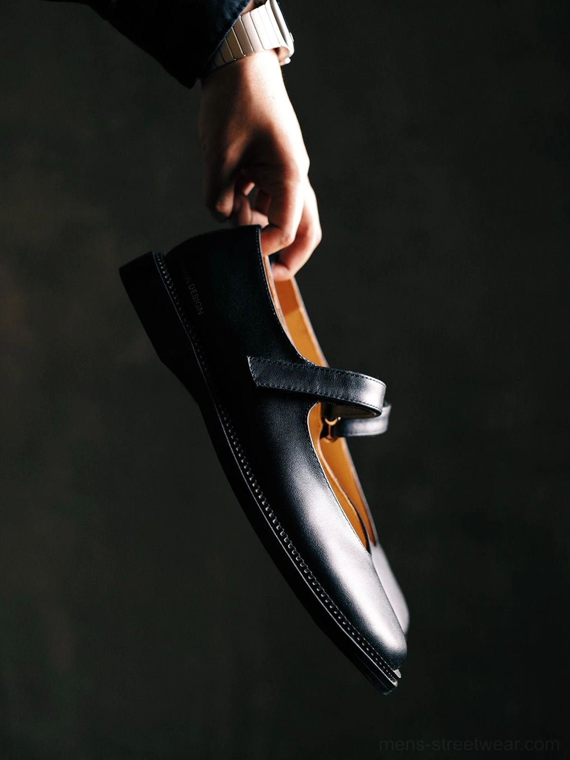 Close-up of a black leather Mary Jane shoe held by the adjustable ankle strap, showing the smooth upper and tan leather-style insole