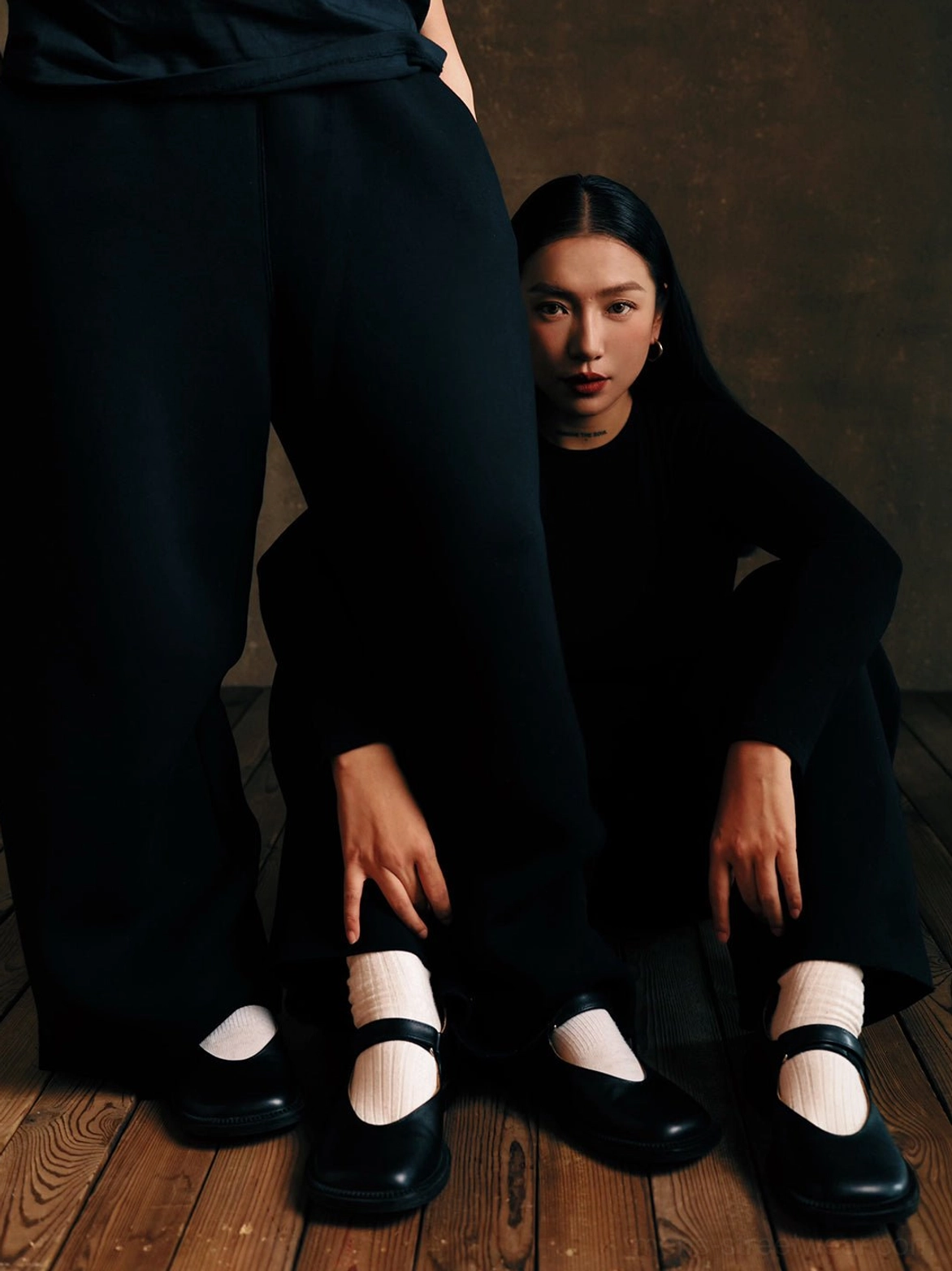 Black leather Mary Jane shoes styled with white socks and matching black outfits in a dark studio with a wood floor and concrete backdrop