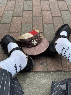 Black leather Mary Jane shoes styled with printed crew socks, pinstripe trousers, and a plaid cap on a brick sidewalk for an East Asian streetwear outfit