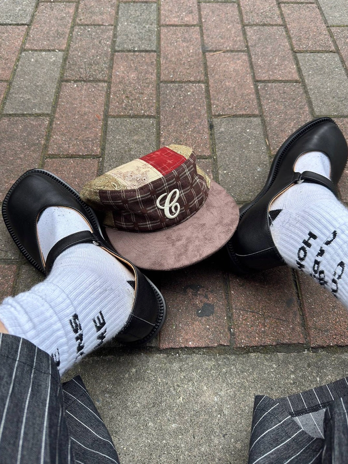 Black leather Mary Jane shoes styled with printed crew socks, pinstripe trousers, and a plaid cap on a brick sidewalk for an East Asian streetwear outfit