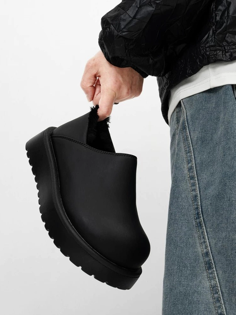 All-black leather platform slip clog held beside loose jeans, highlighting the matte upper, chunky sole, rounded toe, and easy slip-on streetwear silhouette