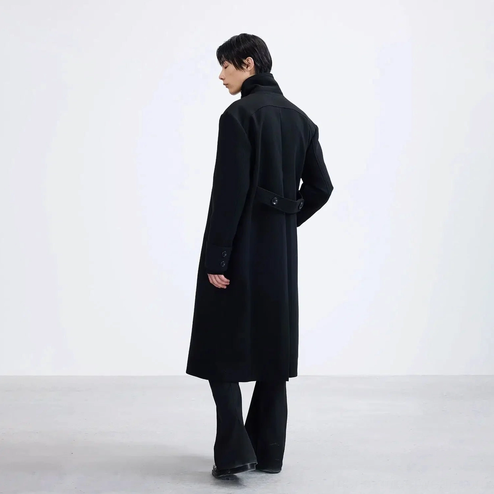 Black long double-breasted overcoat back side view showing adjustable buckle waist belt and straight longline silhouette