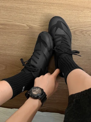 Top-down view of a black low-profile athletic sneaker worn with black crew socks, showing geometric upper panels and tonal flat laces