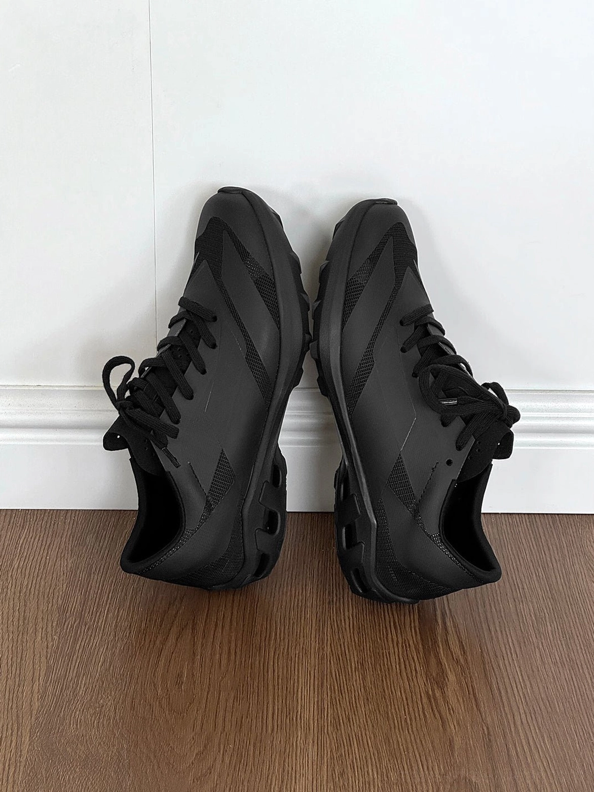 Low-angle side view of black low-profile sneakers on a wooden floor, showing the chunky textured anti-slip outsole and tonal upper