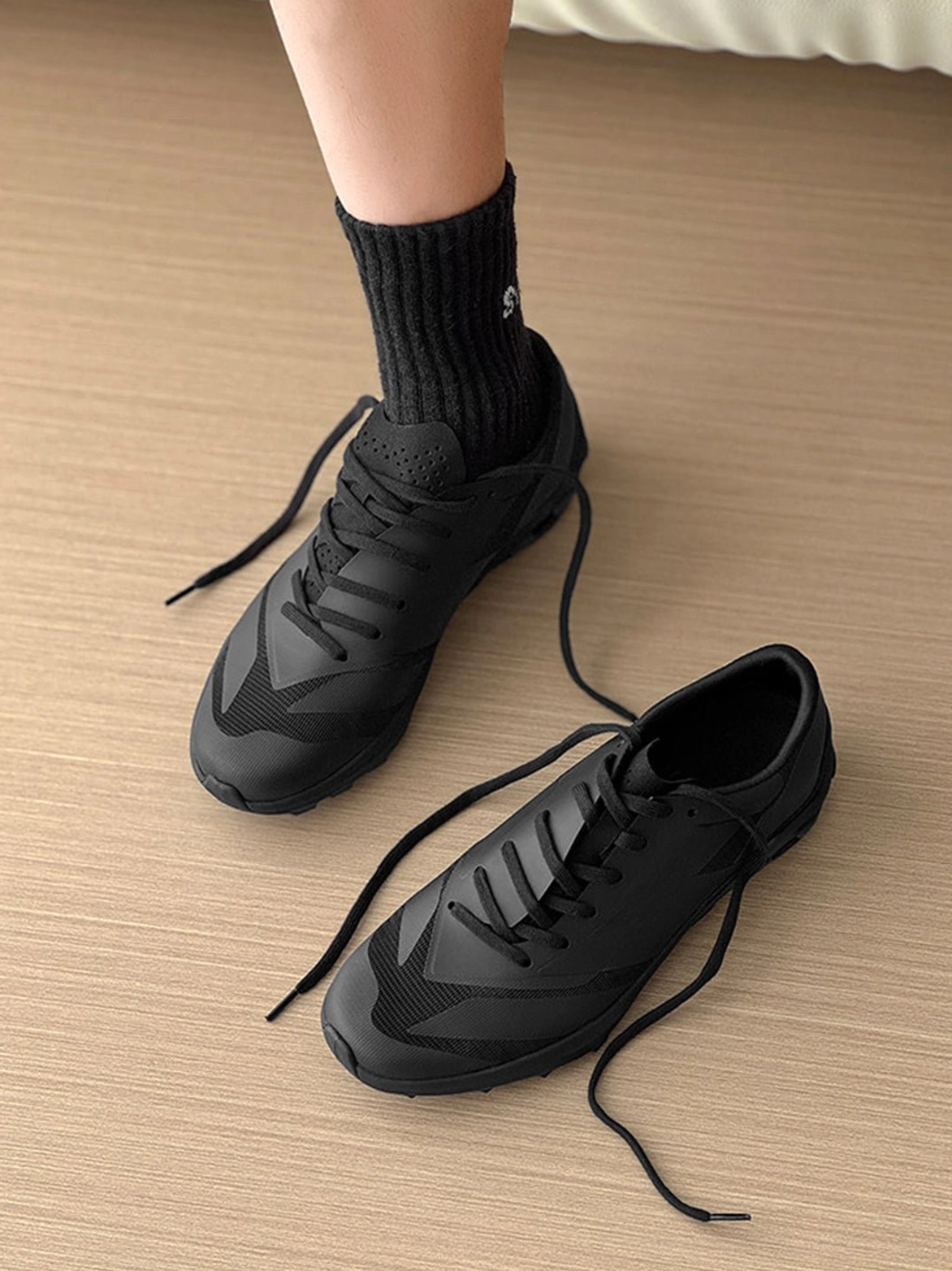 Overhead product view of a black low-profile streetwear sneaker with geometric printed panels, loose black laces, and textured anti-slip outsole