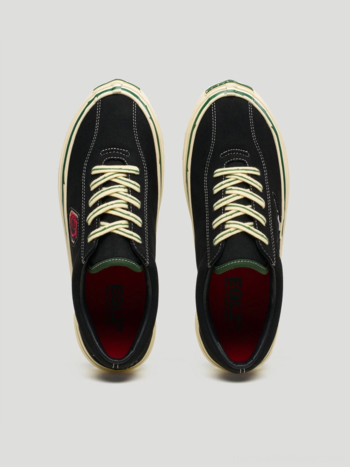 Flat lay of matching black low-top skate shoes with beige laces, red inner lining, and green tongue accents