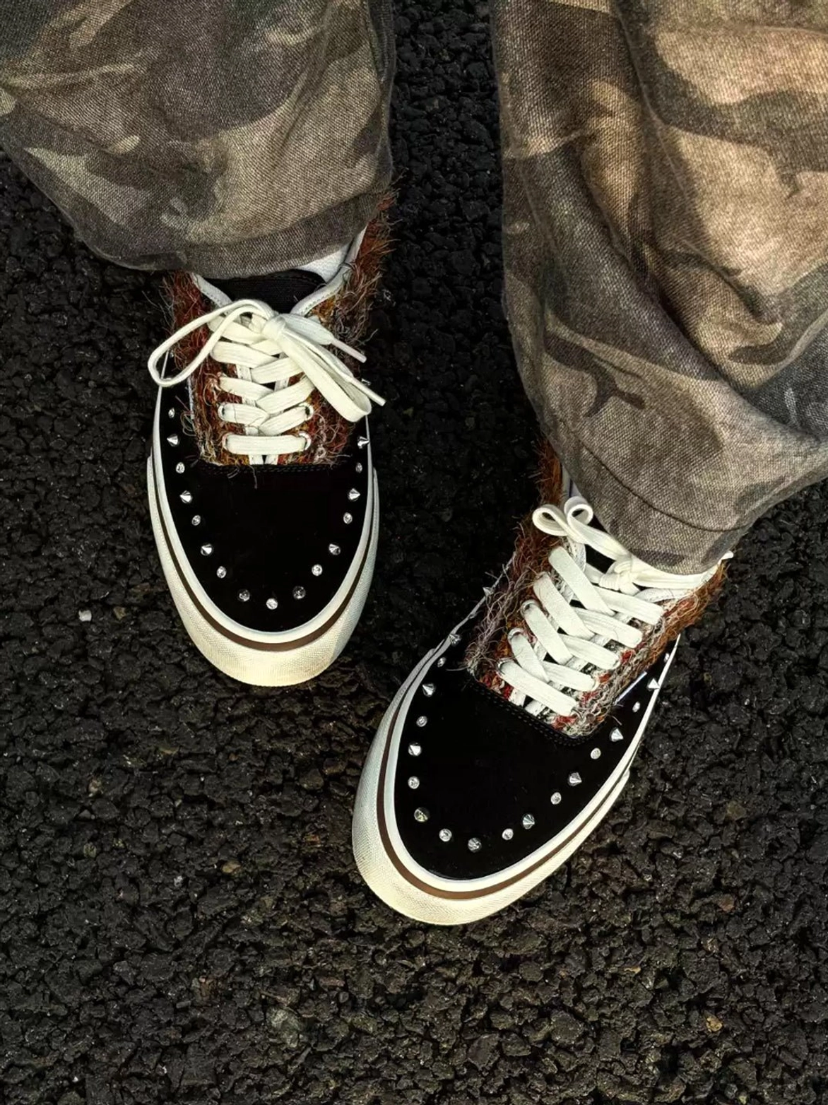 Overhead on-foot view of black low-top studded sneakers with embellished toe caps, brown fuzzy side panels, and white rubber soles