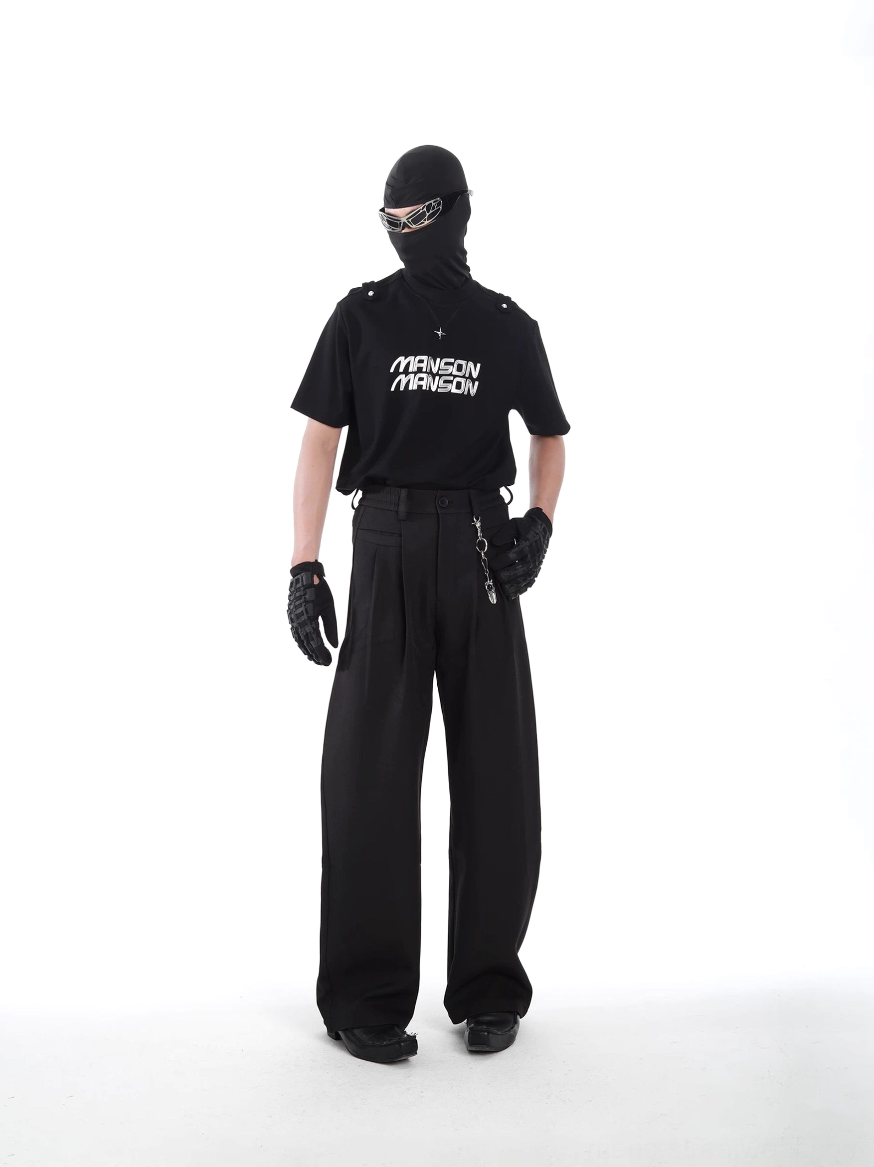 Rear three-quarter styling view of black MANSON graphic streetwear T-shirt with tactical shoulder accents, balaclava, gloves, and black wide-leg pants