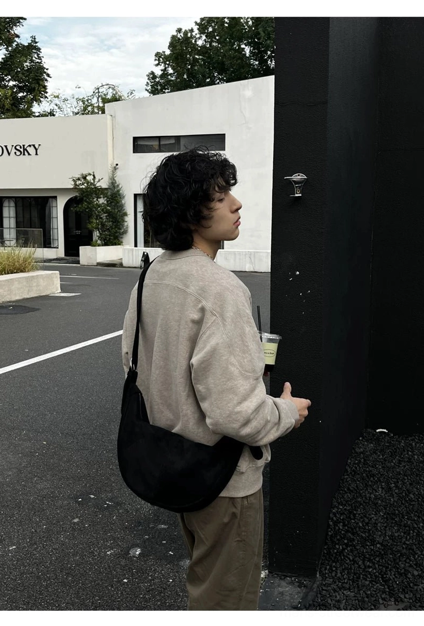 Black matte crescent sling bag worn low at the hip with beige sweatshirt in minimalist Korean streetwear styling