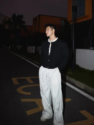 Black men's knit button cardigan layered over a white henley shirt, shown from a back three-quarter view with loose white trousers in a Korean streetwear outfit