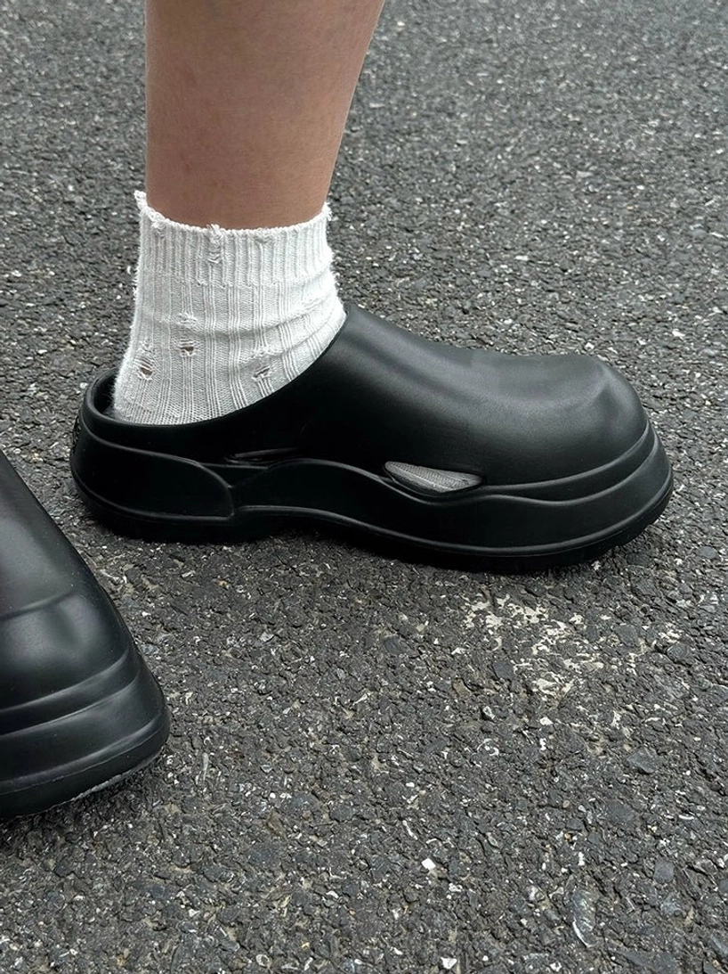 Black men's platform clogs shown from the side on asphalt with distressed white crew socks for a casual urban streetwear outfit