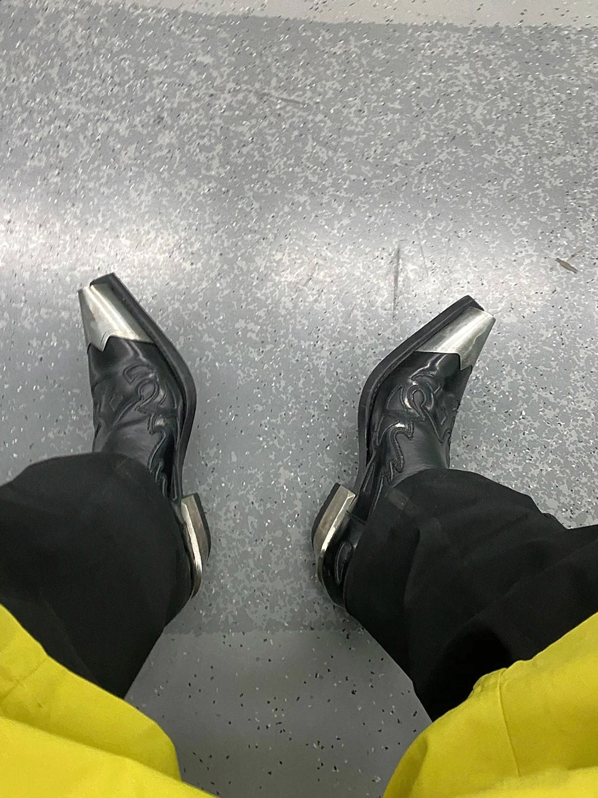 On-foot view of black metallic toe block heel Chelsea boots styled with black trousers and yellow pants on a concrete floor