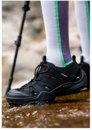 Black outdoor water hiking sandal worn with striped athletic socks on a rocky shallow water trail, showing breathable construction and rugged grip