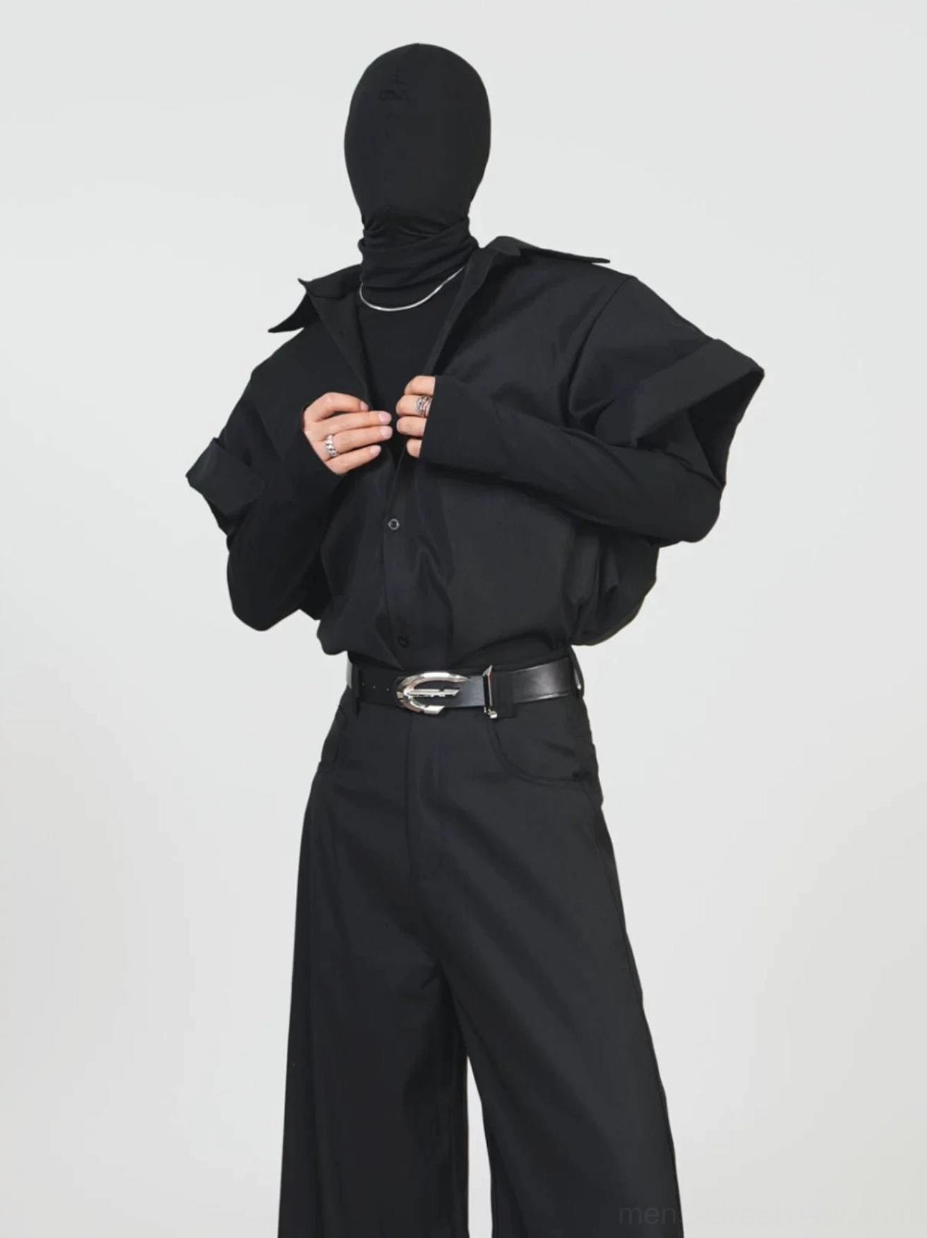 Front upper body view of a masked model adjusting the collar of a black oversized button front shirt jacket over a fitted turtleneck