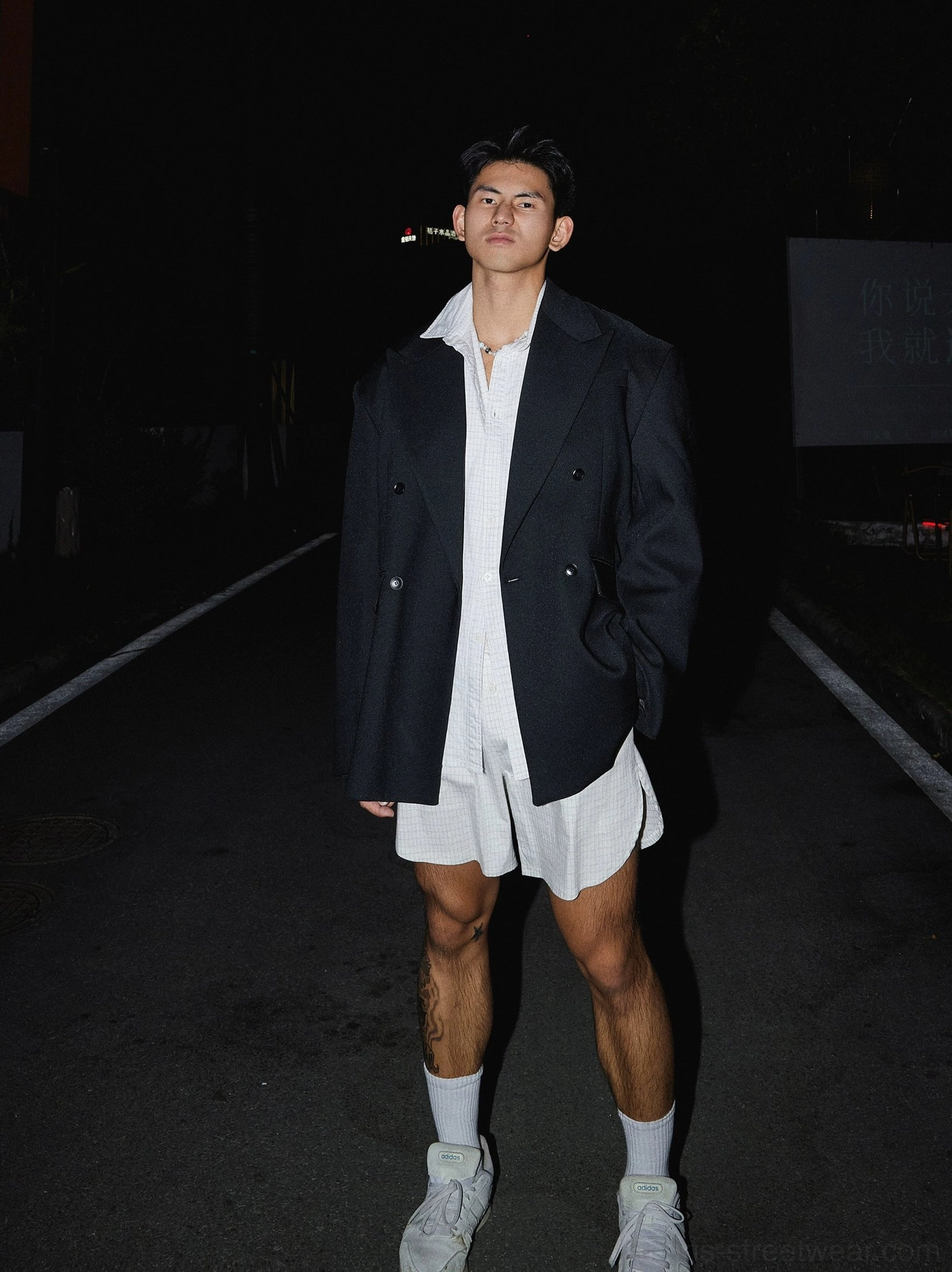 Full-body black oversized double-breasted blazer outfit with grid shirt, shorts, white socks and sneakers on an urban night street