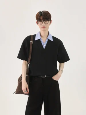 Full-body male model wearing a black oversized faux layered T-shirt with blue striped collar, black wide-leg trousers, glasses, and brown crossbody bag