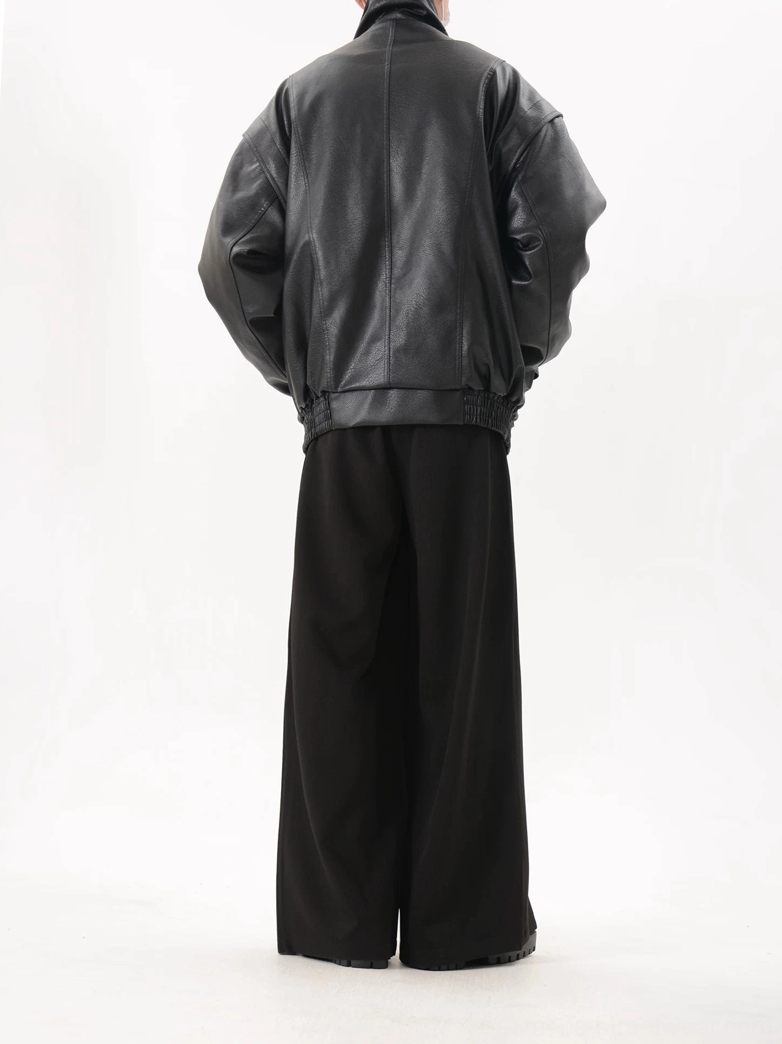 Full-body back view of a black oversized faux leather bomber jacket with dropped shoulder construction, seamless back panel, and gathered elasticated waist hem